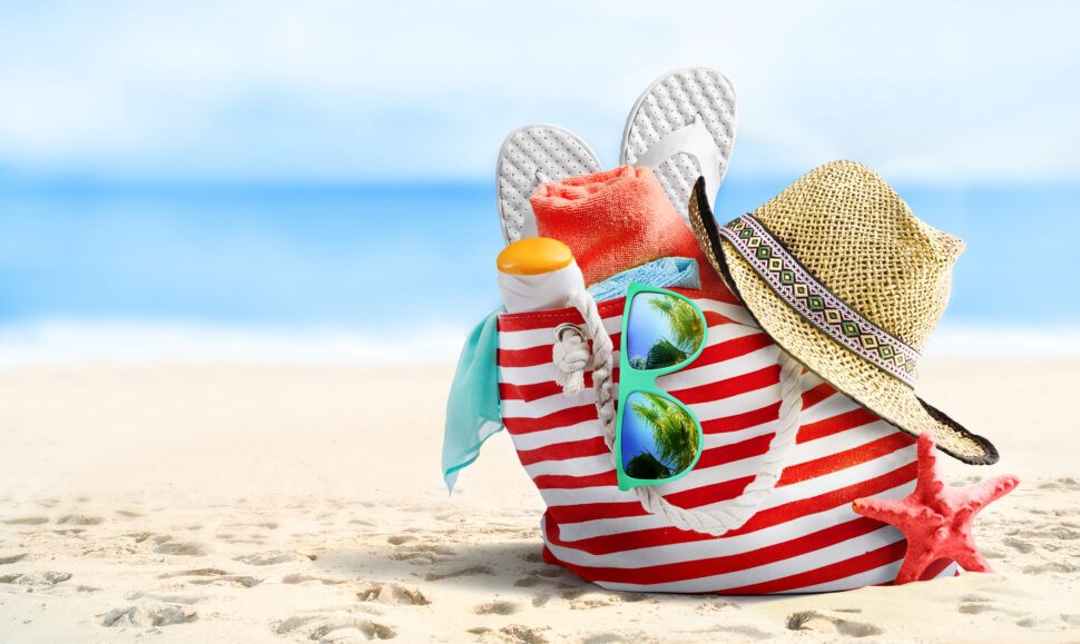 Een strandtas met slippers, zonnebrand, zonnebril, hoed en een handdoek. De tas ligt in het zand op het strand.